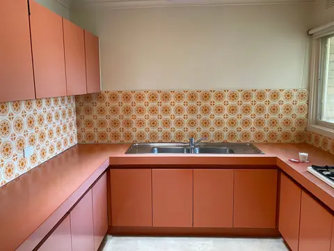 A dated kitchen with salmon-orange cabinets, laminate counters and a retro patterned tile backsplash, lit by a side window